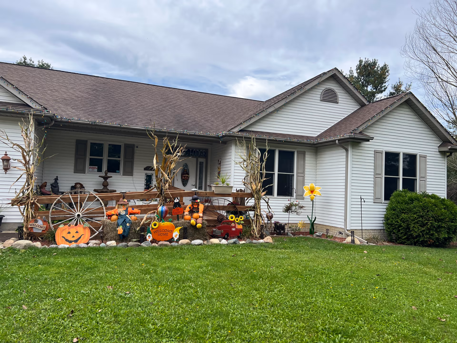 Front exterior view of a single-story white house with a brown roof, decorated with autumn and Halloween-themed items including scarecrows, pumpkins, hay bales, and a wooden wagon wheel. The lawn is green and well-maintained, and there are some bushes and trees around the house.