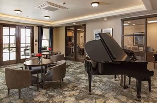 A spacious senior living common area featuring a black grand piano on the right, round tables with chairs arranged for seating, and large windows with glass doors allowing natural light to fill the room. The room has a patterned carpet and neutral-colored walls with ceiling lights.