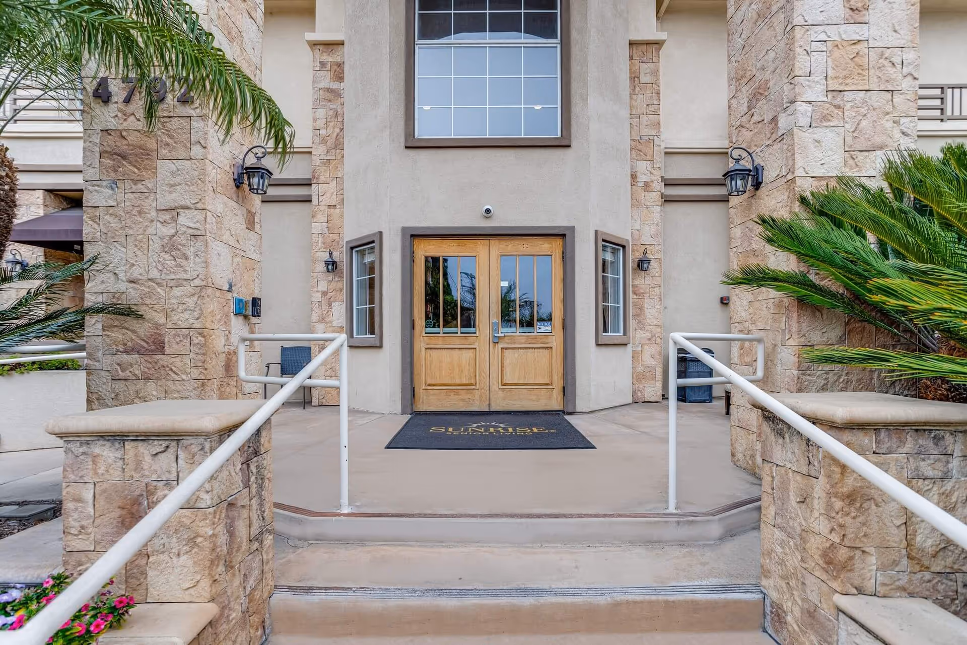 Entrance to a building with stone pillars and beige walls, featuring double wooden doors with glass panels. There are handrails on both sides of the steps leading up to the entrance, and a black mat with the word 'SUNRISE' and a sun logo is placed in front of the doors. Palm plants and other greenery are visible on either side of the entrance.
