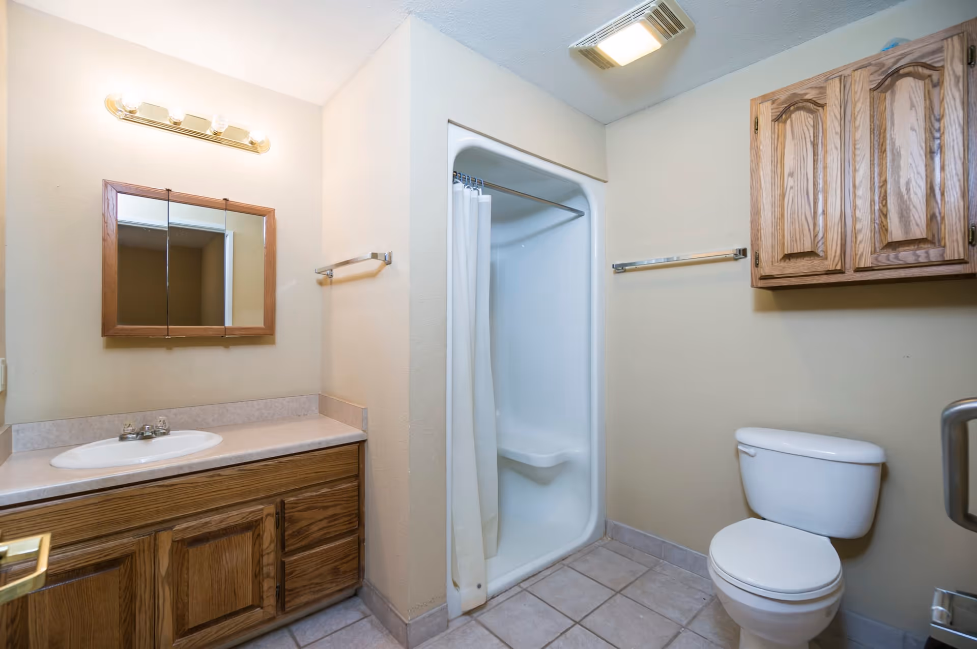 A clean bathroom with a wooden vanity and sink on the left, a mirror with wooden frame above the sink, a shower with a white curtain in the center, and a white toilet with a wooden cabinet mounted on the wall above it on the right. The floor is tiled and the walls are painted beige.