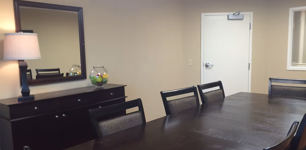 A dining room with a long dark wooden table surrounded by chairs. On the left side, there is a dark wooden sideboard with a lamp and a glass bowl filled with decorative fruit. A large mirror hangs above the sideboard, reflecting part of the room. The walls are beige, and there is a white door in the background.