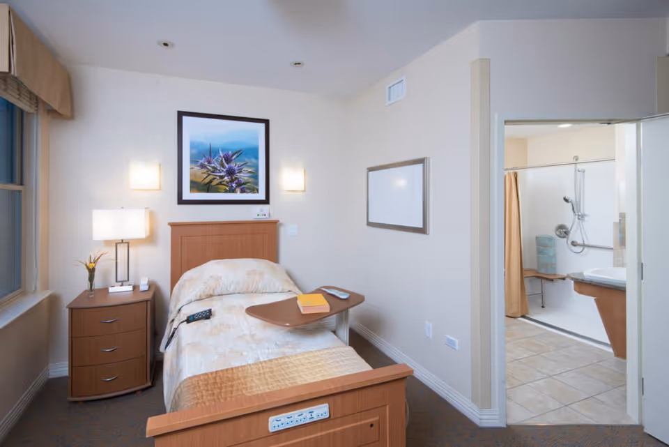 A single bed with a beige and light brown bedspread in a small room. Next to the bed is a wooden nightstand with a lamp, a small plant, and a phone. Above the bed is a framed picture of a purple flower with a blue sky background. On the right wall is a whiteboard. An open door reveals a bathroom with a shower, grab bars, and a beige curtain. The room has a window with a beige valance and carpeted floor.