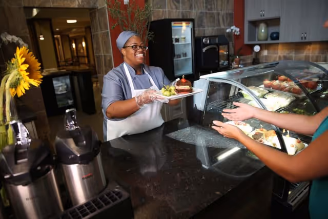 A smiling cafeteria worker wearing a hairnet, glasses, and gloves serves a plate of food including grapes and a dessert to a person across the counter in a food service area with a display case and coffee dispensers.