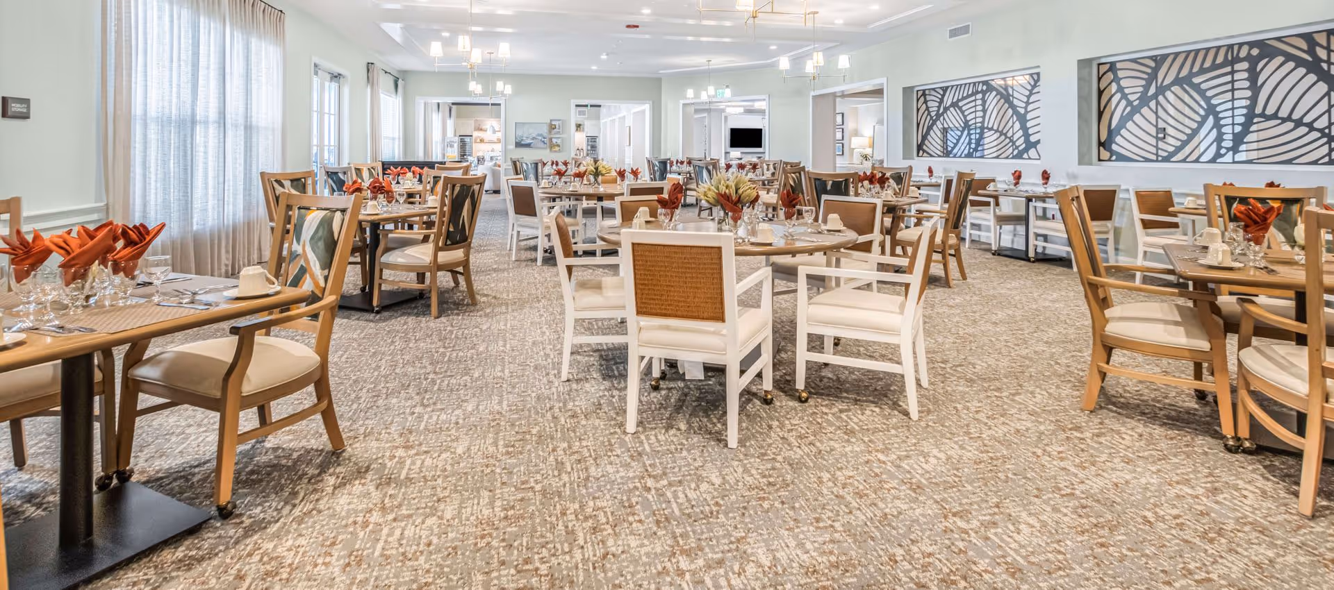 A spacious dining room with multiple tables set with glasses, cups, and folded red napkins. The room has light-colored walls, large windows with sheer curtains, and decorative wall art. The chairs are a mix of wooden and cushioned styles, and the carpet has a patterned design.