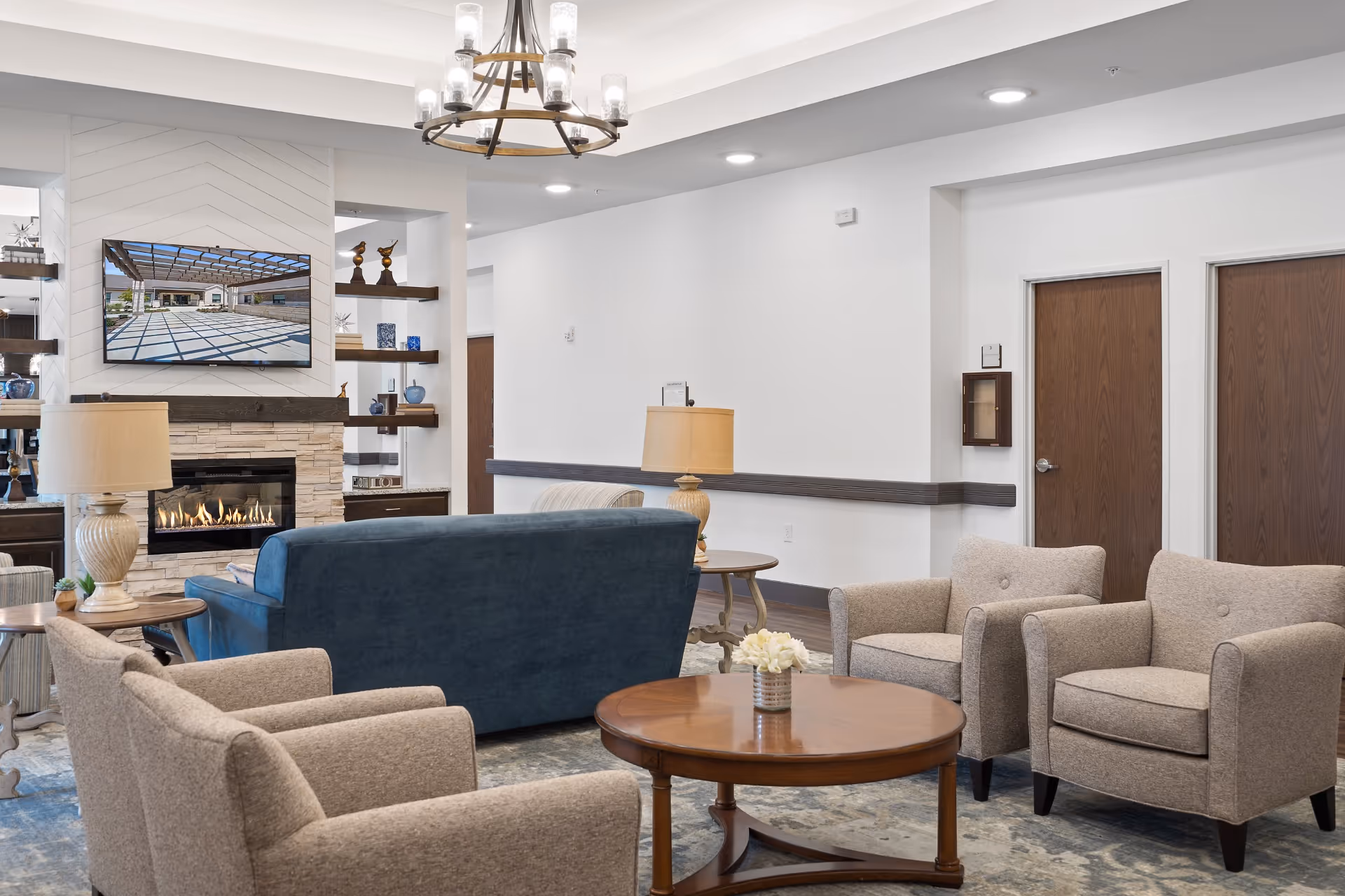 A cozy lobby area featuring a blue sofa, beige armchairs, a wooden coffee table with a floral centerpiece, and a stone fireplace. A large TV is mounted on the wall, surrounded by decorative shelves.