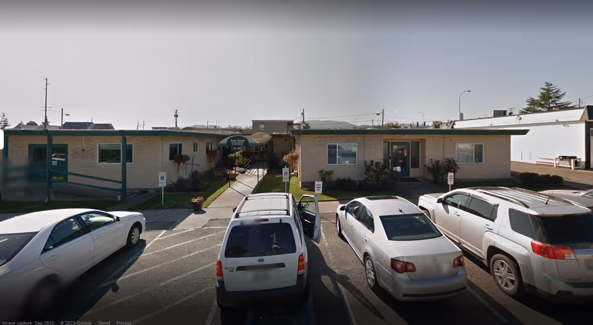 Exterior view of a single-story assisted living facility building with a green awning entrance labeled 'San Juan'. Several cars are parked in front of the building in a parking lot with designated handicapped parking spaces.