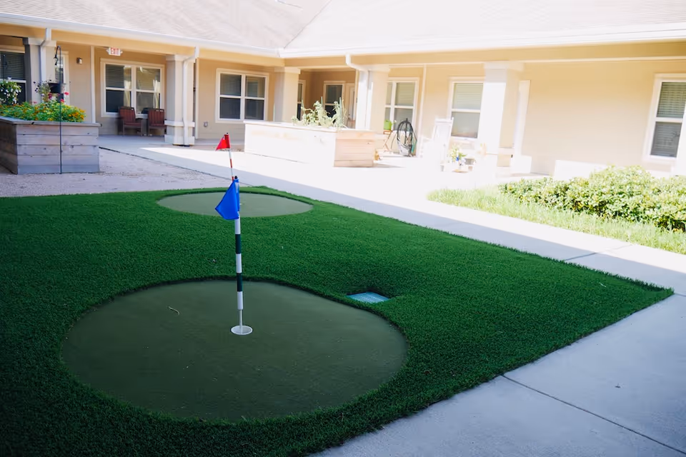 Small outdoor putting green with two holes and flags in a courtyard in front of a senior living building.