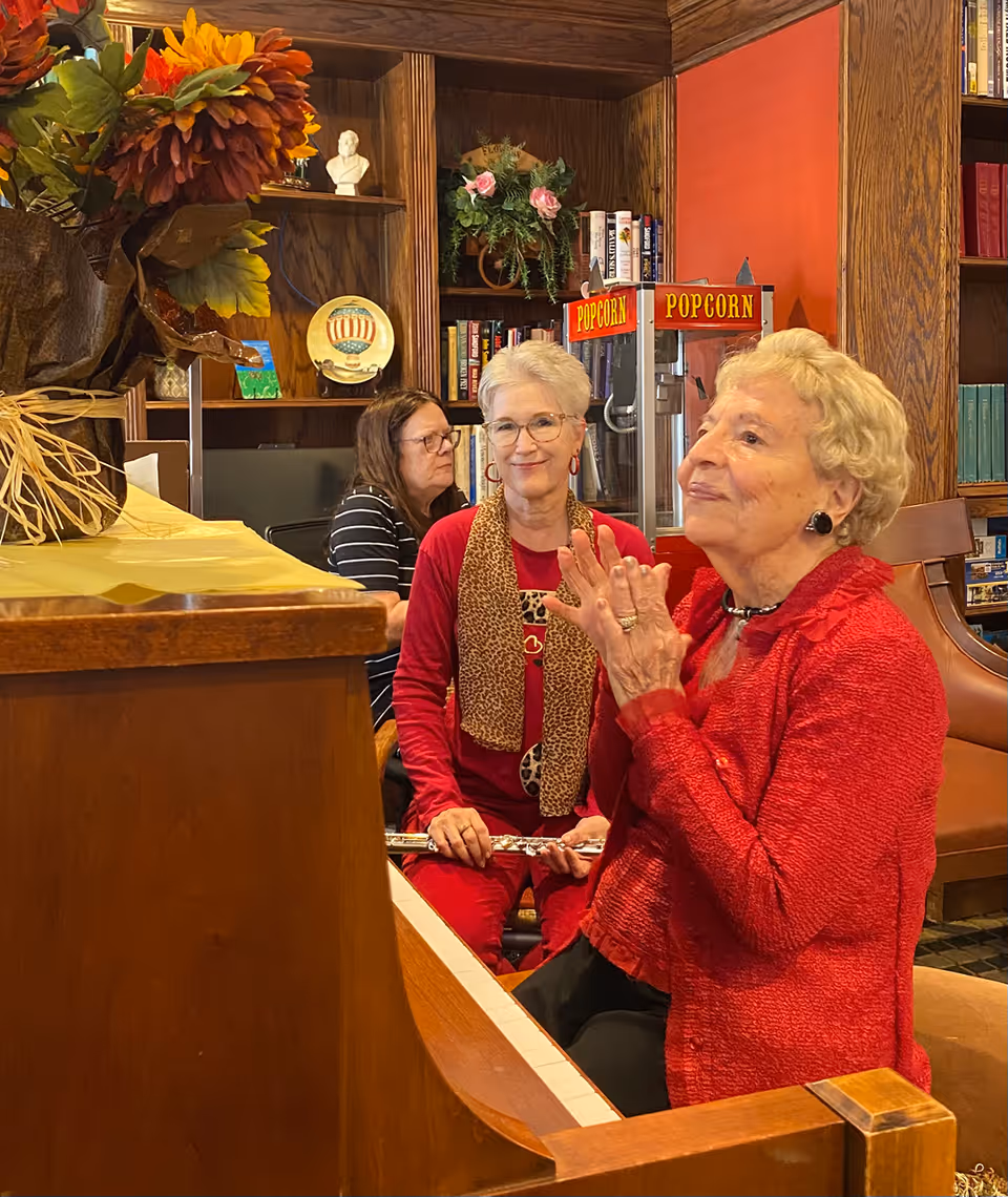Three older women in a library-style common room, one seated at a piano and another clapping, with bookshelves and a popcorn machine behind them.