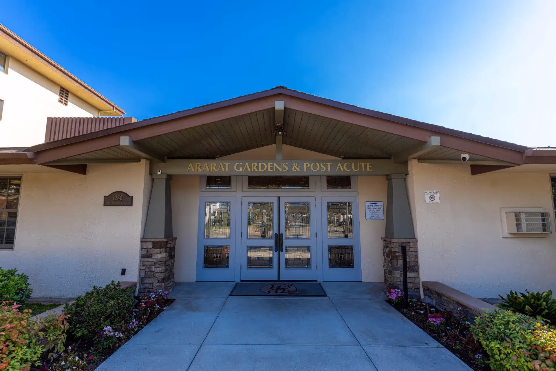 Front entrance of Ararat Gardens & Post Acute facility with double glass doors, a covered porch with a peaked roof, and landscaped flower beds on either side of the walkway.