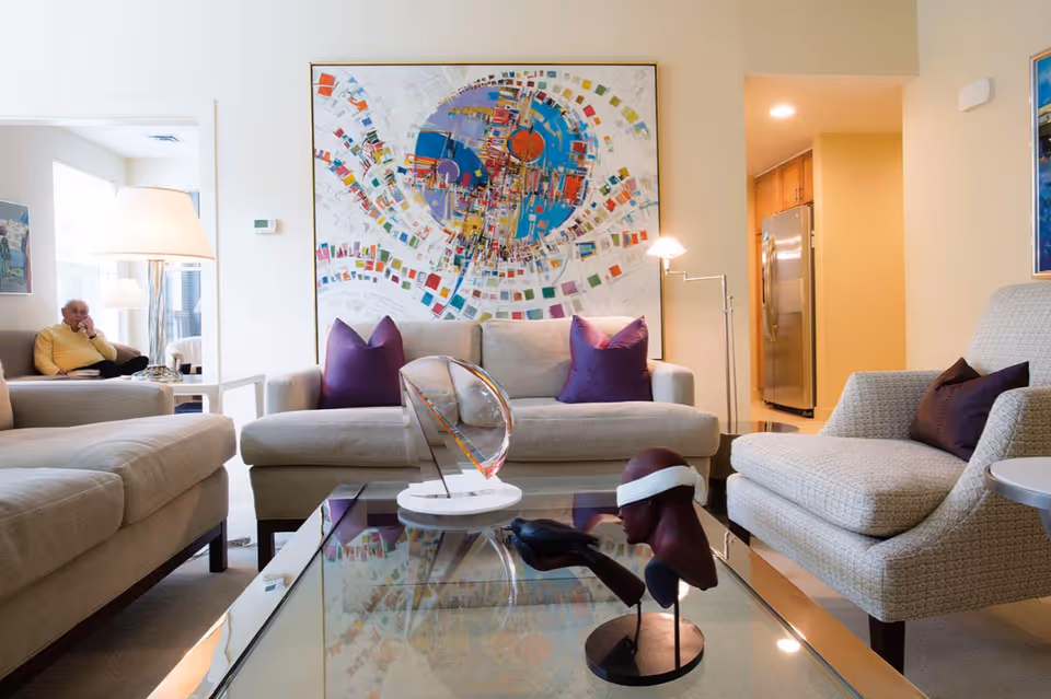 A modern living room with beige sofas and purple pillows arranged around a glass coffee table displaying two artistic sculptures. A large colorful abstract painting hangs on the wall behind the sofa. In the background, an elderly man in a yellow sweater is sitting in a separate room near a window with a lamp beside him. The kitchen area with wooden cabinets and a stainless steel refrigerator is visible through an open doorway.