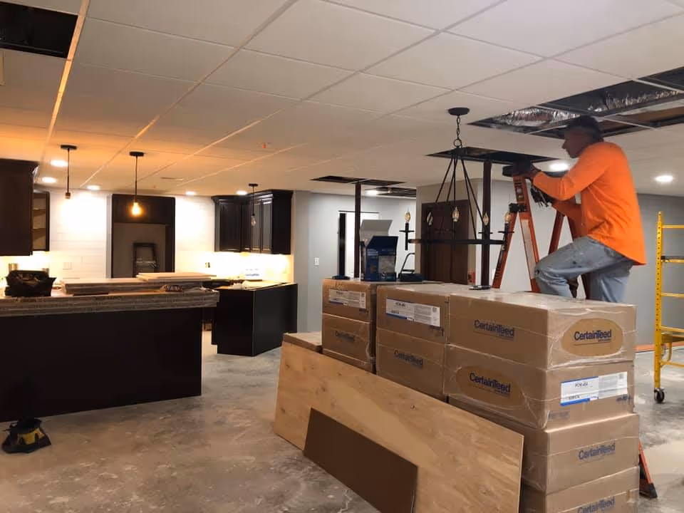 A man in an orange shirt and blue jeans is standing on a ladder working on the ceiling in a room under construction or renovation. The room has dark kitchen cabinets, a granite countertop, and several boxes labeled 'CertainTeed' stacked on a wooden platform. The ceiling has some missing tiles, and construction materials and tools are visible around the space.
