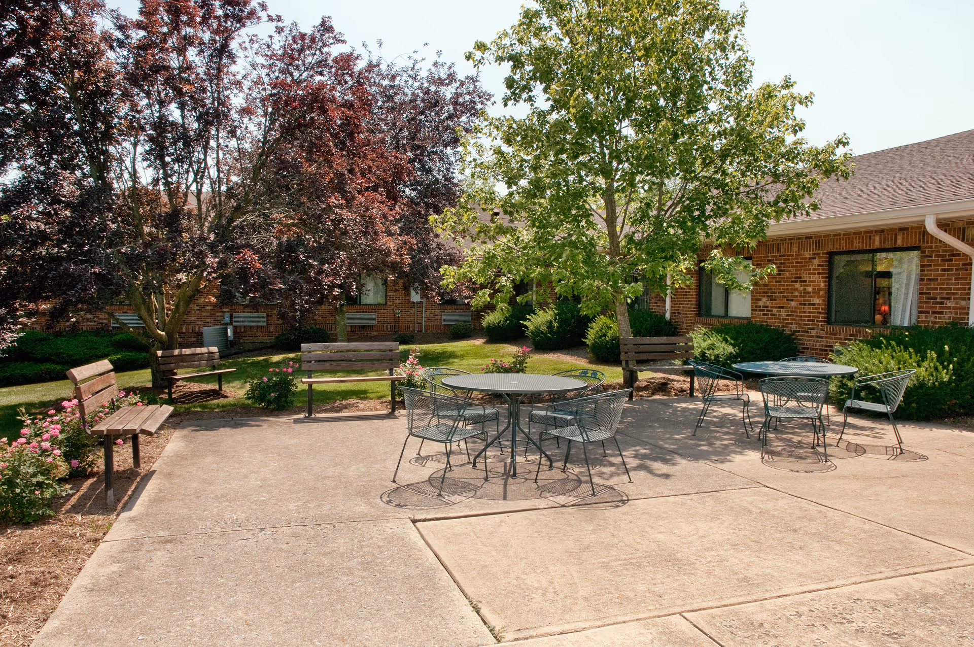 Outdoor patio courtyard with metal tables and chairs, wooden benches, trees, and a brick building in the background.
