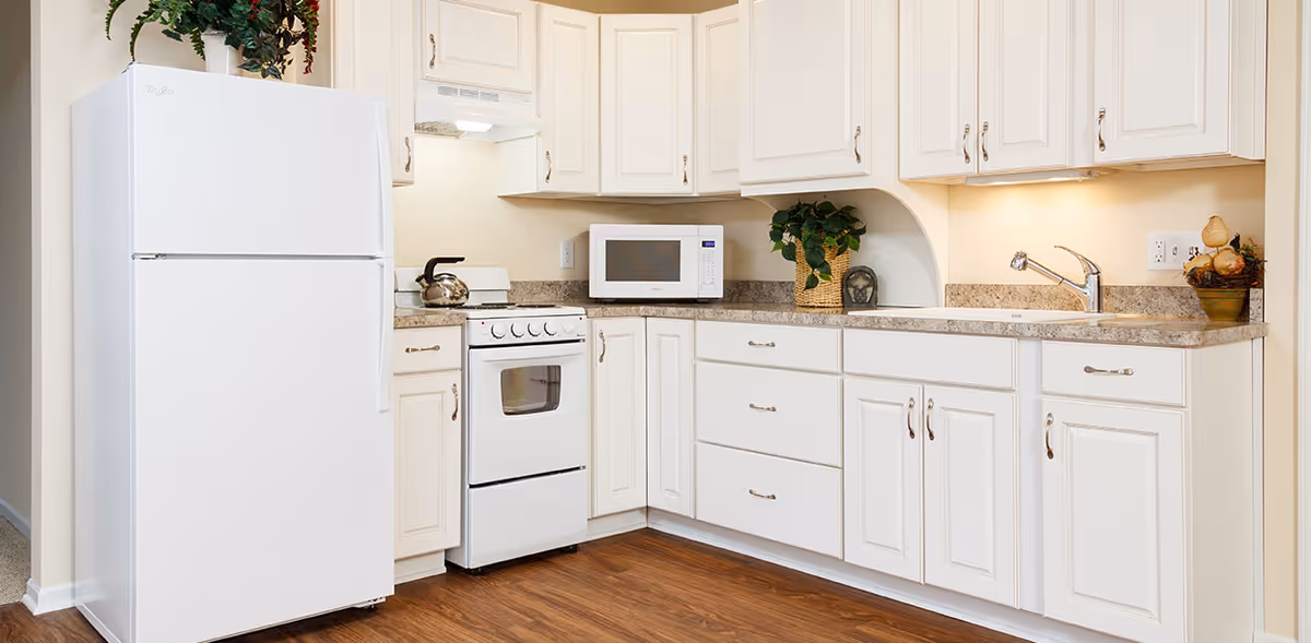A clean and bright kitchen featuring white cabinets, a white refrigerator, a white stove with a kettle on top, a microwave on the countertop, a sink with a faucet, and decorative plants placed on the counter and above the refrigerator. The floor is wooden and the walls are painted a light beige color.