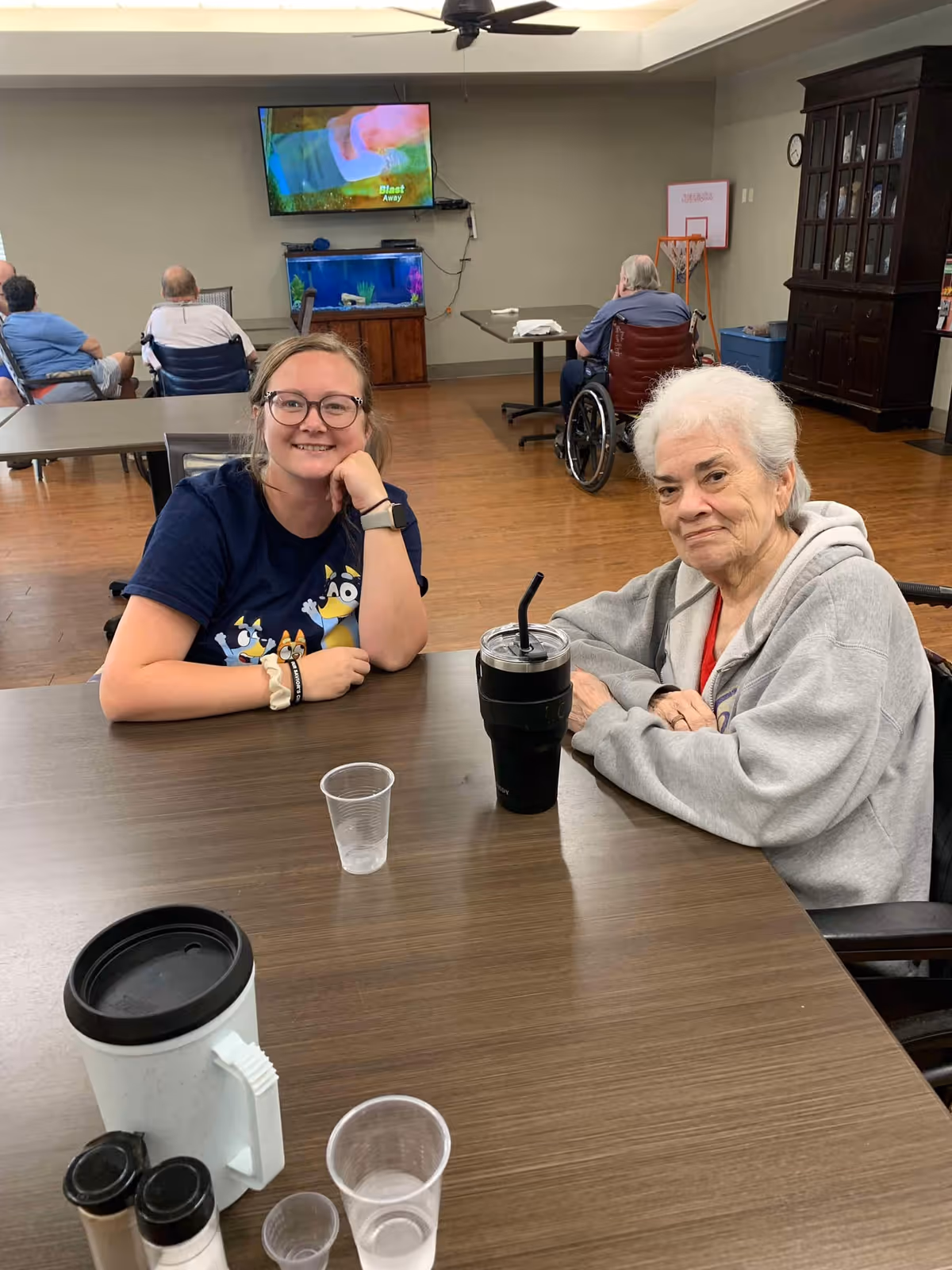 Two women sitting at a table in a common area of a senior living facility. One woman is younger with glasses and a dark blue shirt, smiling with her hand resting on her chin. The other woman is older with white hair, wearing a gray hoodie, and sitting in a wheelchair. On the table are a large black tumbler with a straw, a white mug with a black lid, and several clear plastic cups. In the background, other residents are seated, a television is mounted on the wall above a fish tank, and a small basketball hoop is visible.