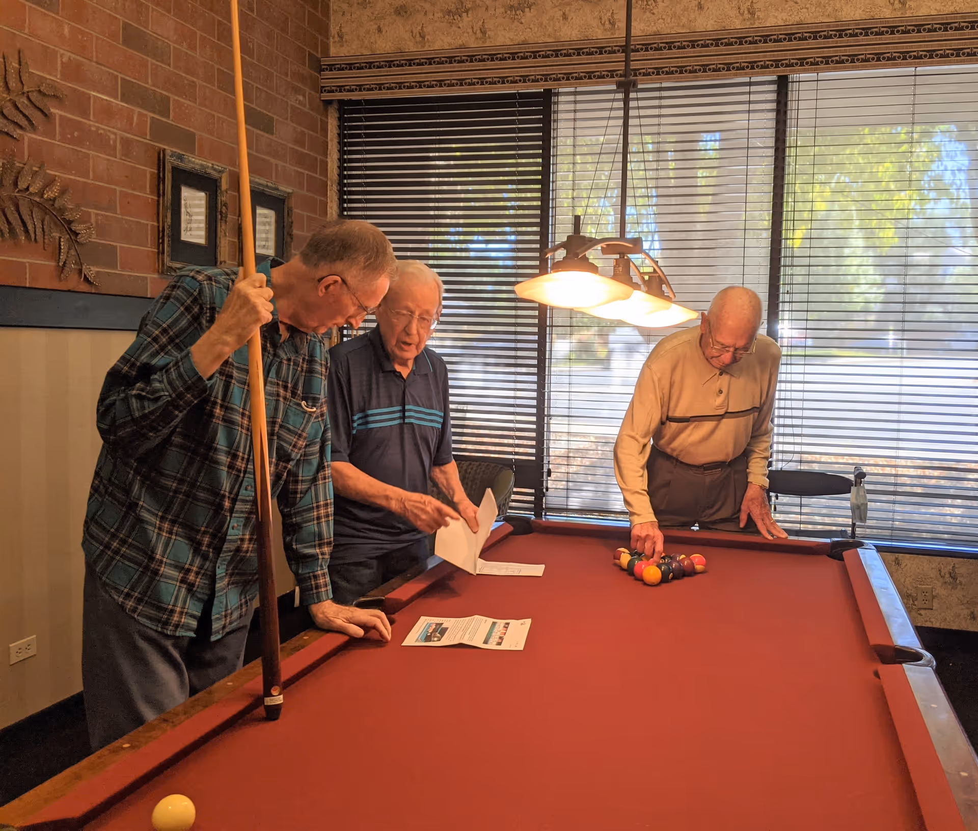 Three elderly men standing around a red pool table indoors. One man is holding a pool cue, another is holding papers, and the third is arranging billiard balls in a triangle rack. The room has large windows with blinds and a hanging light fixture above the pool table.