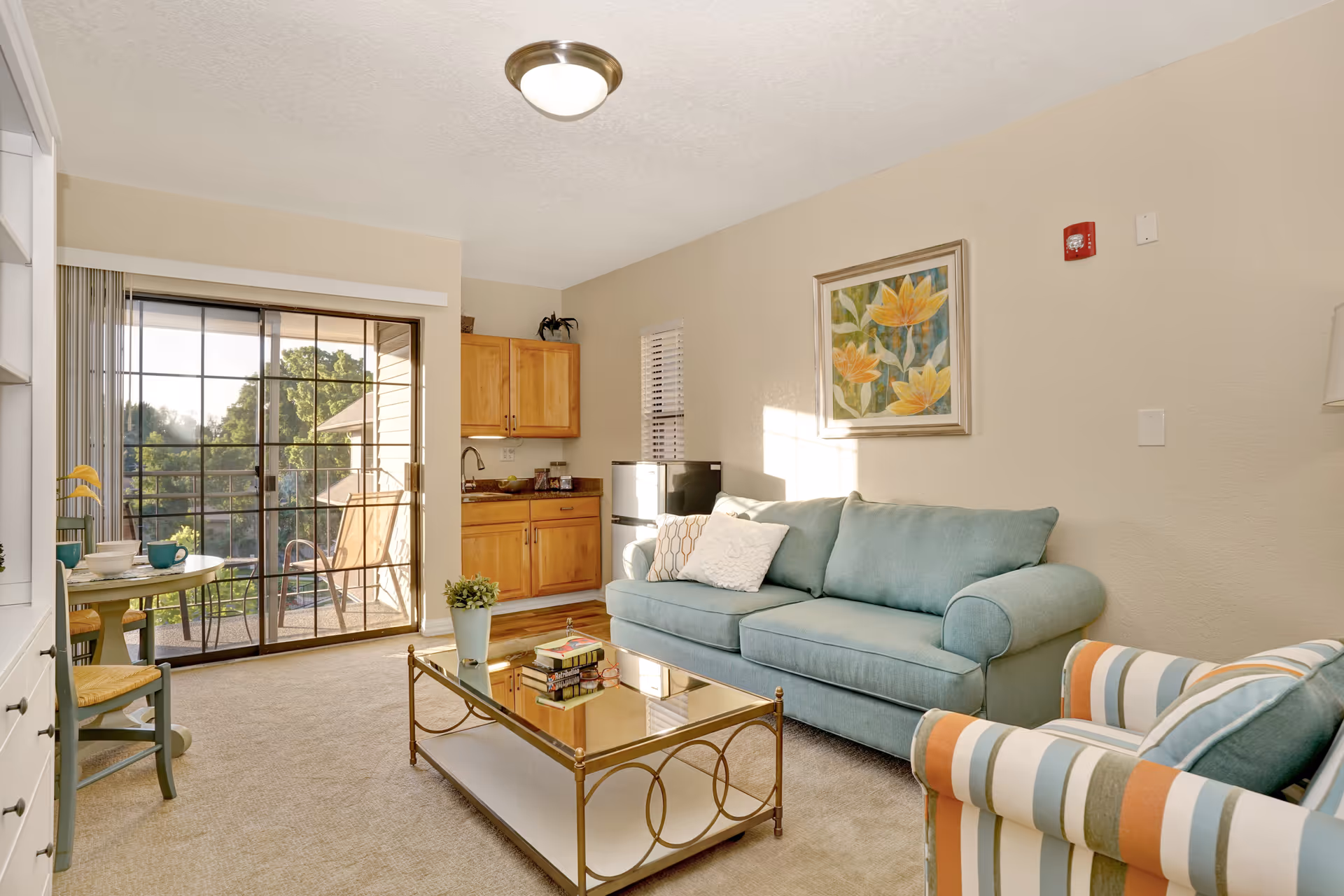 A bright living room with a teal sofa, a striped armchair, a glass coffee table with books and a plant, and a small kitchenette with wooden cabinets and a mini fridge. There is a sliding glass door leading to a balcony with outdoor chairs and a view of trees outside. A round dining table with two chairs is partially visible on the left.