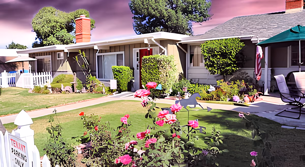 Exterior view of a single-story senior care facility with a well-maintained garden featuring pink roses, trimmed bushes, a white picket fence, and outdoor seating under a green umbrella. The building has a red door and a chimney, with a purple sky in the background.