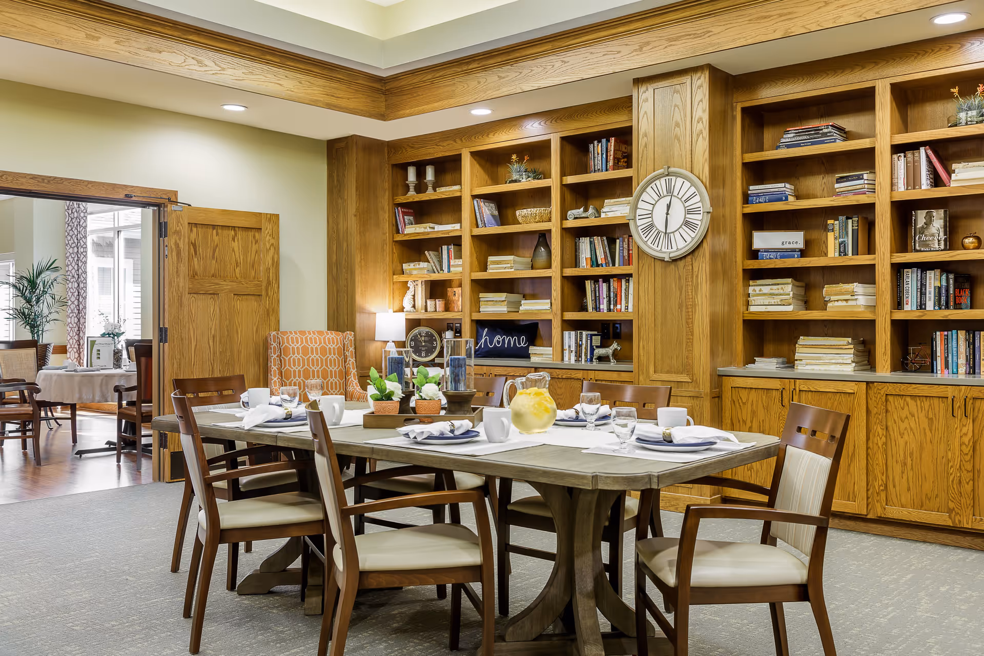 A cozy dining area with a wooden table set for six people, featuring white plates, cups, and a pitcher of lemonade. The room has built-in wooden bookshelves filled with books and decorative items, a large wall clock, and soft lighting. Adjacent to the dining area is another room with additional seating and a potted plant near a window.
