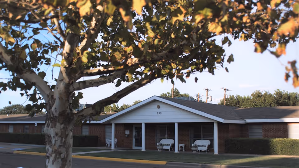 Single-story brick senior care building with a covered entrance marked "630", white columns, porch chairs, and a tree in the foreground.