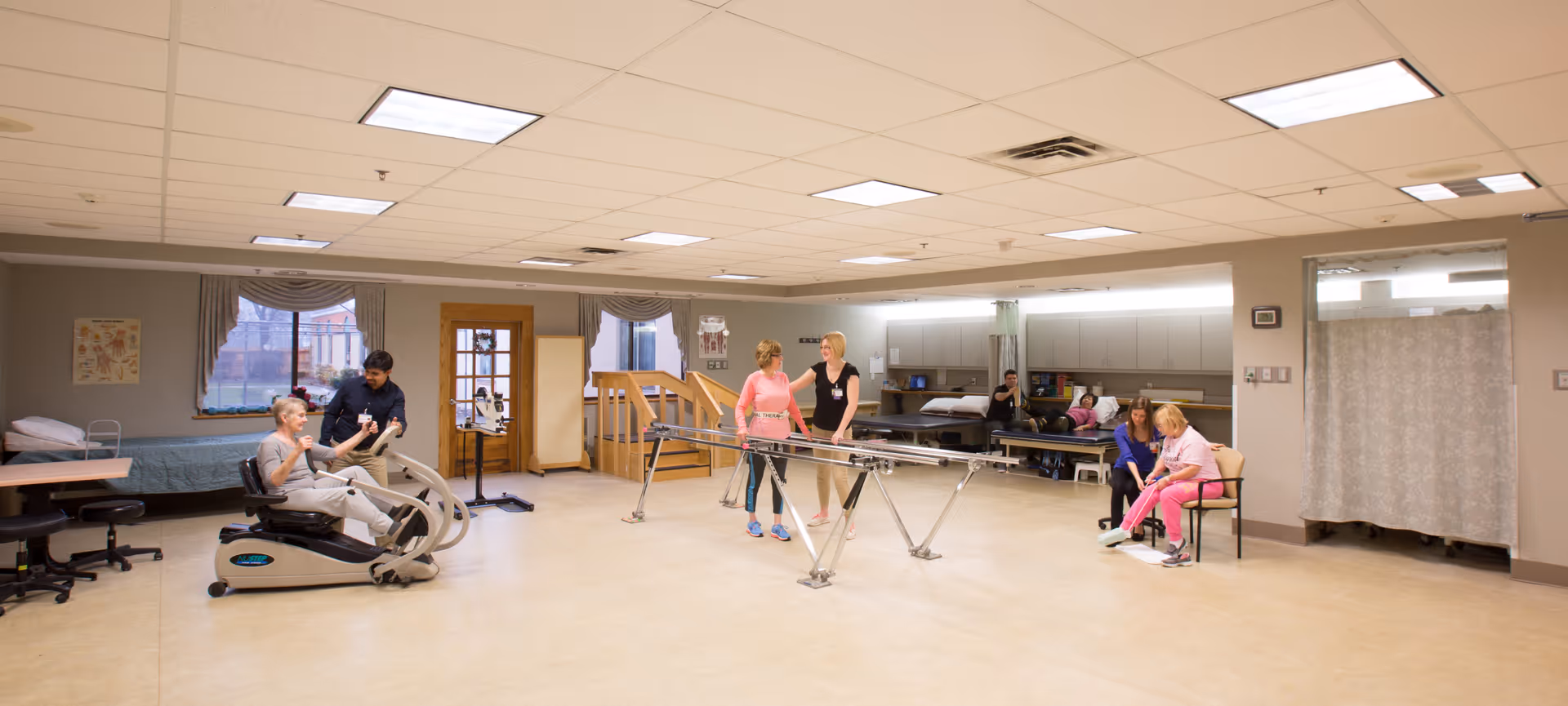 A spacious rehabilitation room where seniors use exercise equipment and parallel bars while staff assist and observe.