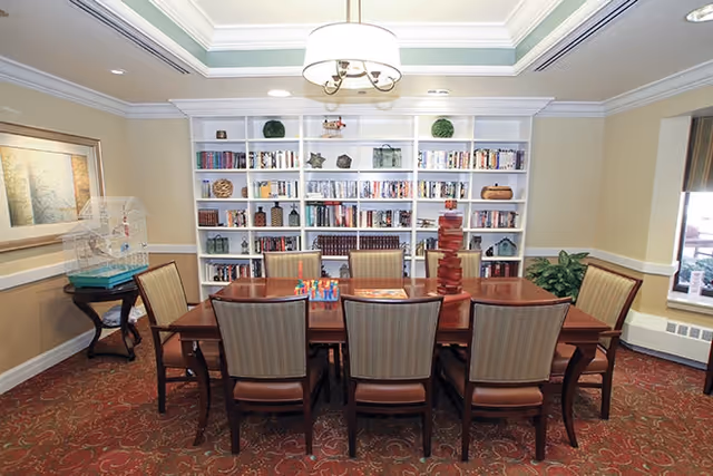 A communal activity room with a long wooden table surrounded by chairs and a large built-in bookshelf filled with books and decorative items under a ceiling light.