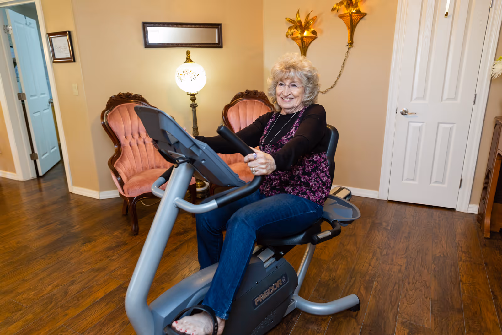 An elderly woman with gray hair and glasses is smiling while sitting on a stationary exercise bike in a room with wooden floors. Behind her are two vintage pink upholstered chairs, a decorative lamp, and a closed white door.