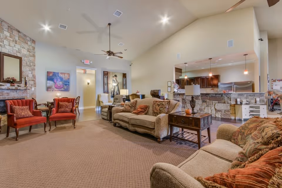 A spacious and warmly decorated living room area in a senior living facility with beige walls and carpet. The room features comfortable upholstered sofas and armchairs with decorative pillows, two red armchairs near a stone fireplace with a mirror above it, a wooden side table with a lamp, ceiling fans, and pendant lights hanging over a stone bar counter that opens to a kitchen area with dark wood cabinets and stainless steel appliances.