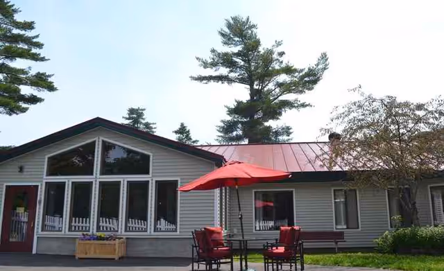 Single-story community building exterior with large windows, a red metal roof, and outdoor seating with a red umbrella.
