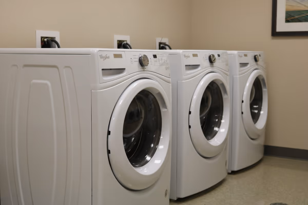 A row of three white front-loading washing machines in a laundry room with beige walls and a framed picture on the wall.