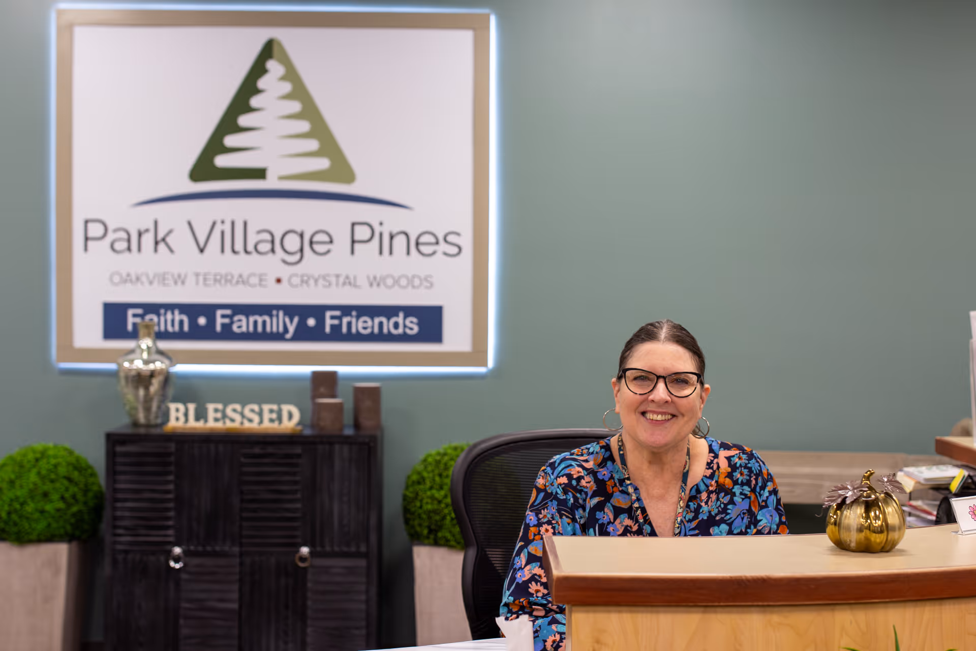 A smiling woman with glasses sitting behind a reception desk in a senior living facility. Behind her is a sign that reads 'Park Village Pines' with the tagline 'Faith • Family • Friends'. The background includes decorative plants and a cabinet with the word 'BLESSED' displayed on top.