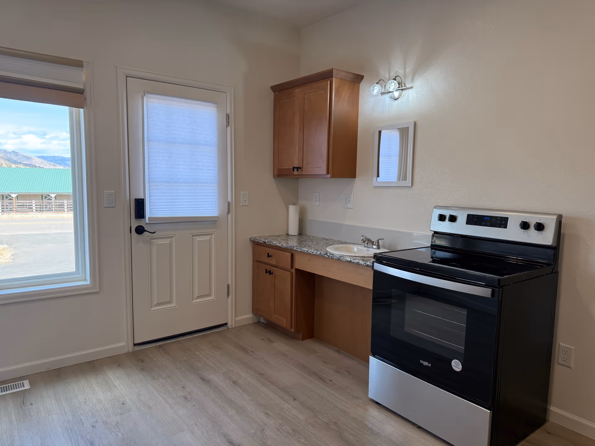 A small kitchen area with a stainless steel electric stove, wooden cabinets, a granite countertop with a built-in sink, and a wall-mounted light fixture above a small mirror. There is a white door with a window covered by a blind, and a large window next to the door showing an outdoor view with a green-roofed building and mountains in the background.