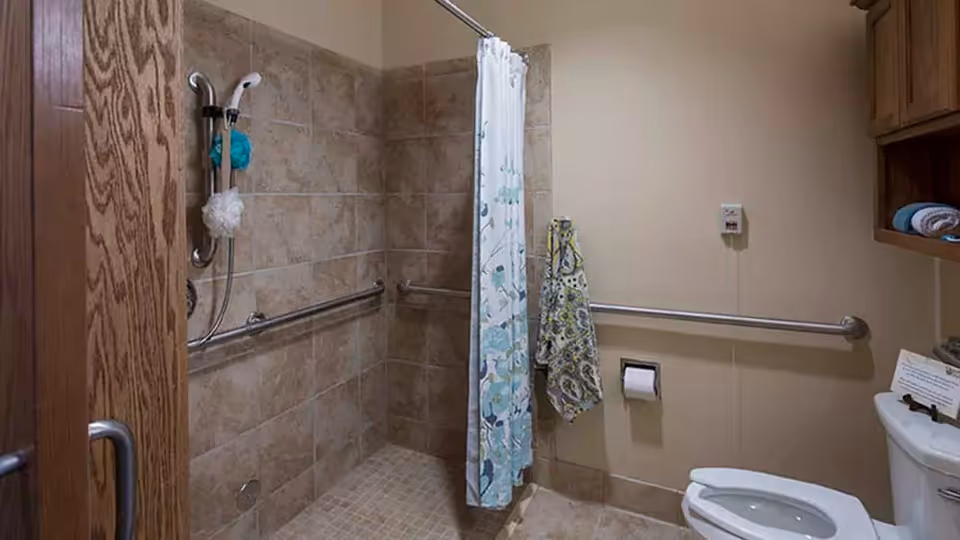 Bathroom with beige tiled walls and floor, featuring a walk-in shower with a handheld showerhead and grab bars. A floral shower curtain partially covers the shower area. There is a toilet with a grab bar on the wall beside it, a toilet paper holder, and a wooden cabinet mounted on the wall with towels inside.