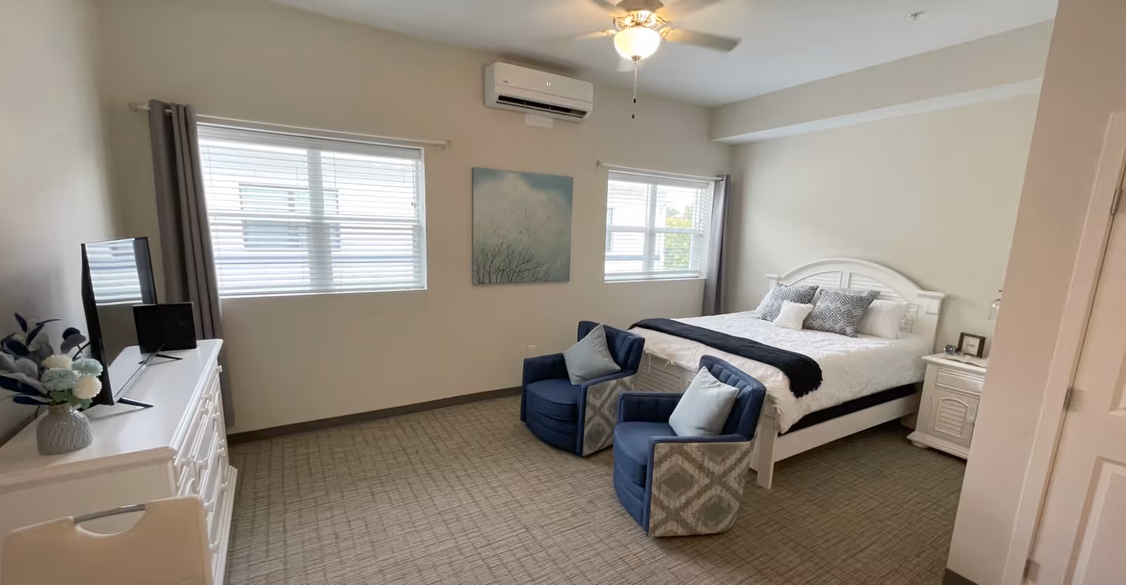 Bright furnished bedroom with a bed, two blue armchairs, a dresser with TV, and two windows.