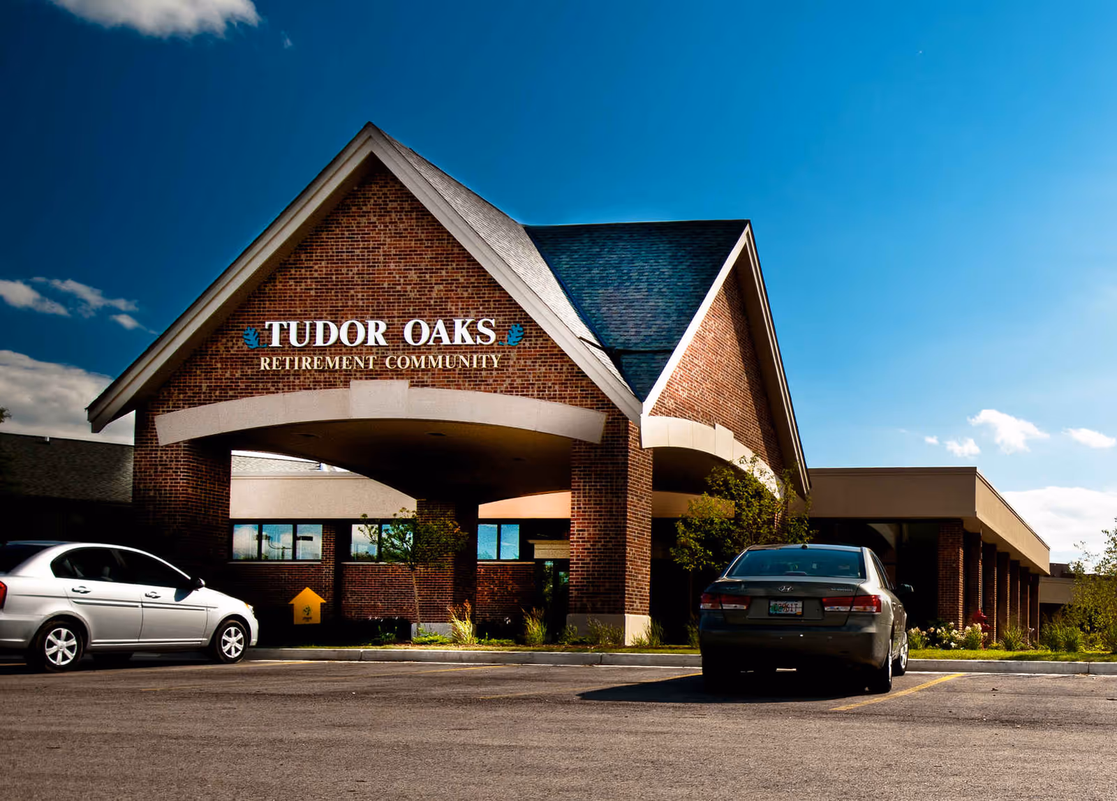 Exterior view of Tudor Oaks Retirement Community building with a brick facade and a peaked roof under a blue sky. Two cars are parked in front of the entrance.