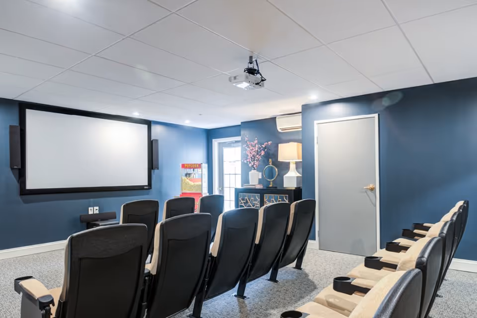 A small movie theater room with two rows of cushioned seats facing a large blank projector screen mounted on a blue wall. There is a popcorn machine in the corner near a door, a cabinet with decorative items and a lamp, and a ceiling-mounted projector above the seats.