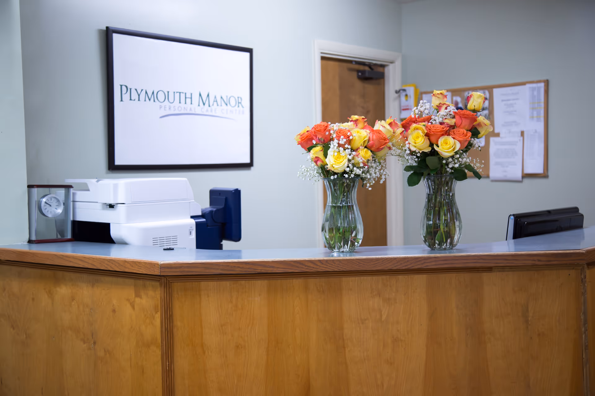Reception desk area at Plymouth Manor Personal Care Center with two glass vases holding bouquets of yellow and orange roses, a printer, a small clock, a bulletin board with papers, and a door in the background.