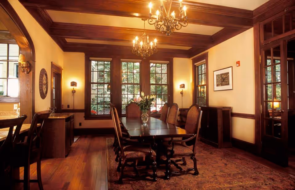 A warmly lit dining room with a long wooden table surrounded by eight upholstered chairs. The room features large windows with wooden frames, wooden ceiling beams, two chandeliers, wall sconces, and a patterned rug on the wooden floor. There is a vase with flowers on the table and a sideboard against the wall.