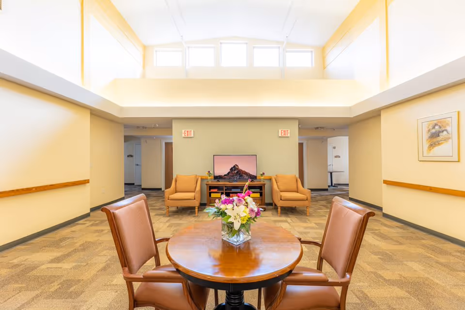 A bright and spacious common area with high ceilings and windows near the ceiling letting in natural light. In the foreground, there is a round wooden table with a vase of colorful flowers and four brown cushioned chairs. In the background, two beige armchairs are placed on either side of a TV on a wooden stand. The walls are painted light yellow and beige, and there are exit signs above two doorways behind the armchairs. A framed picture hangs on the right wall.