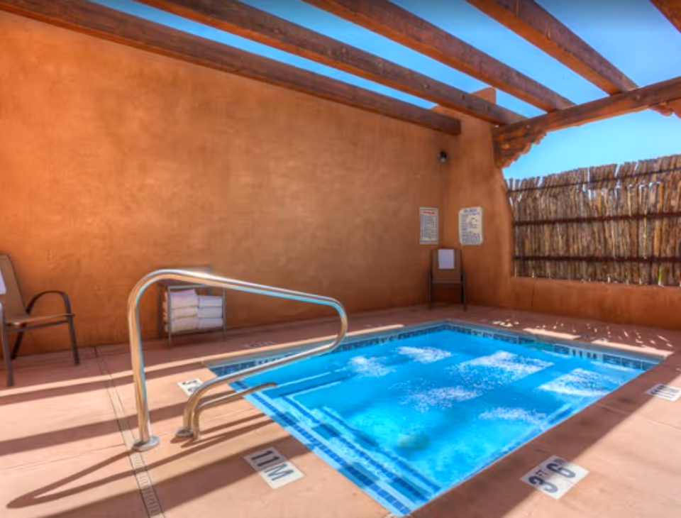 Small outdoor hot tub with bubbling blue water under a wooden pergola and adobe walls.