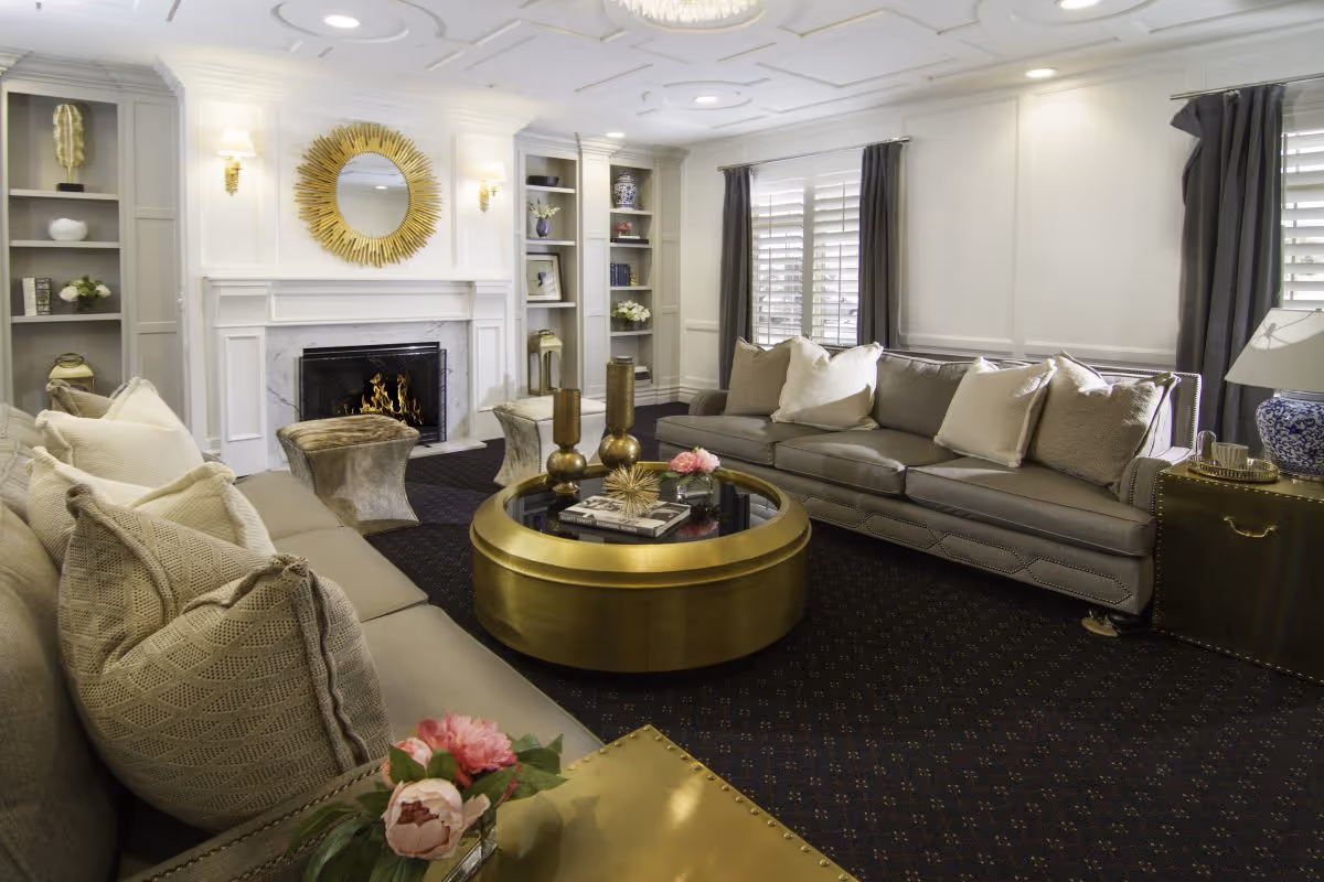 A cozy and elegant living room with two gray sofas adorned with beige and white pillows facing each other around a round gold coffee table. The room features a white fireplace with a decorative gold sunburst mirror above it, built-in shelves with decorative items, dark curtains on windows with white shutters, and a dark patterned carpet. There are also two small upholstered stools and a side table with a blue and white lamp and a tray with cups.