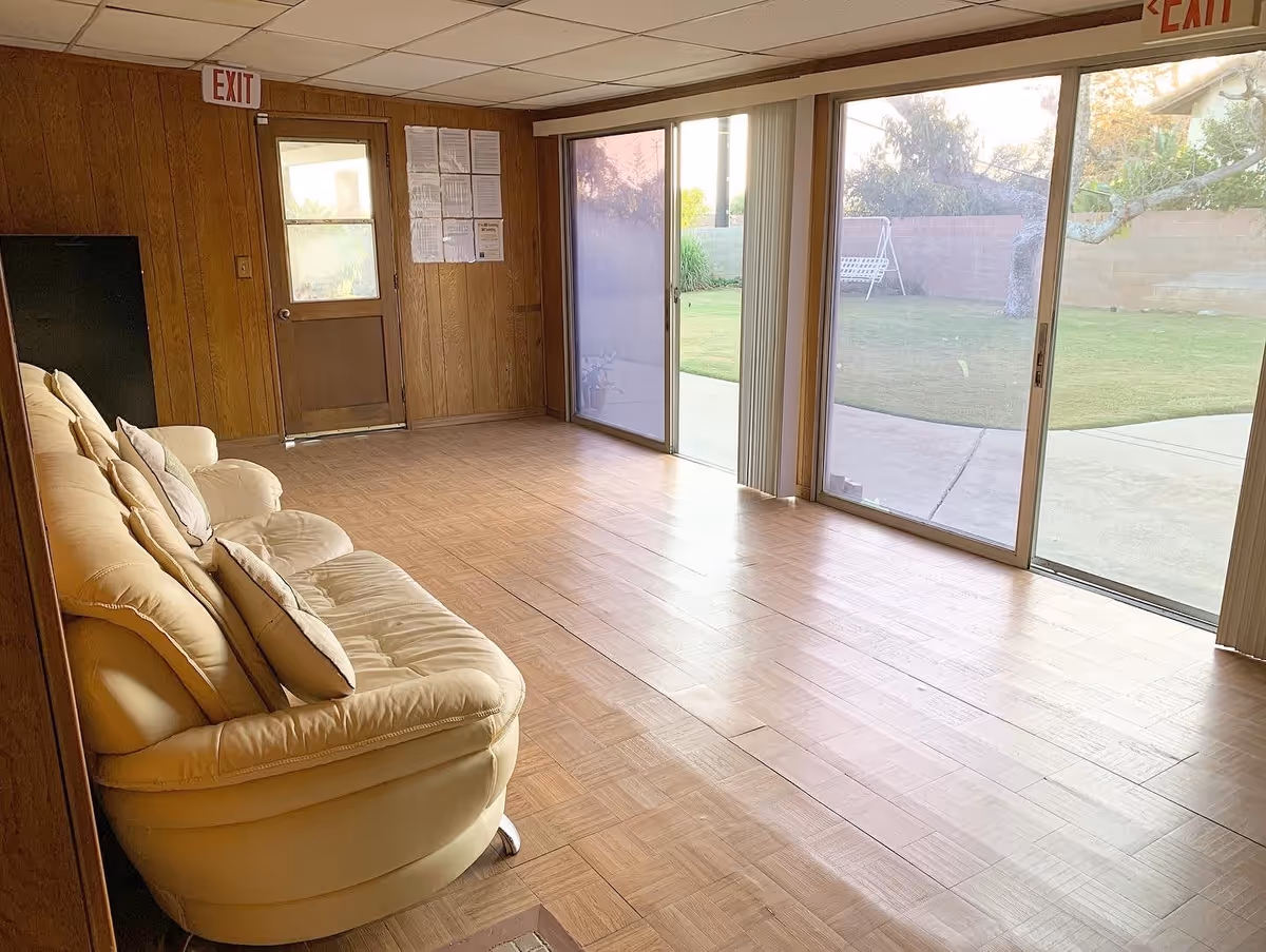 A living room with a beige leather sofa with cushions on the left side, wood-paneled walls, a door with an exit sign above it, and large sliding glass doors on the right side that open to a grassy outdoor area with a swing bench.