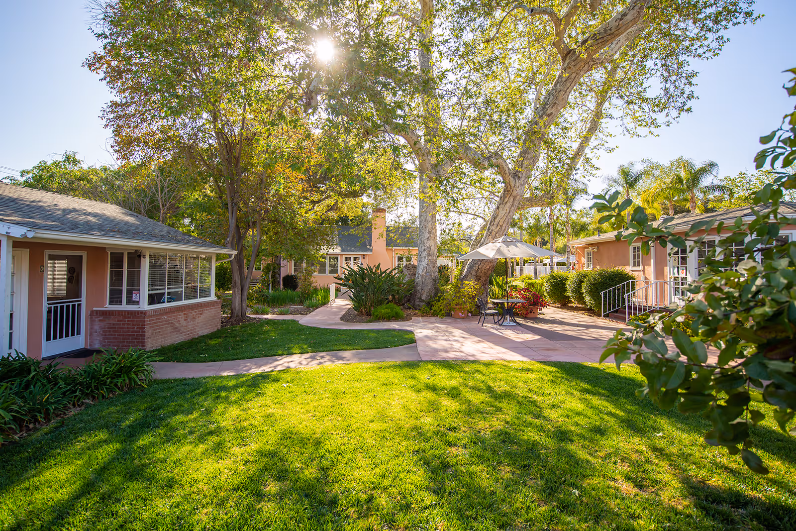 Sunny outdoor courtyard area at Gables of Ojai with green grass, large trees, and a paved walkway. There are several small buildings with pinkish walls and white trim surrounding the courtyard. A table with chairs and an umbrella is set up on the paved area under the trees.