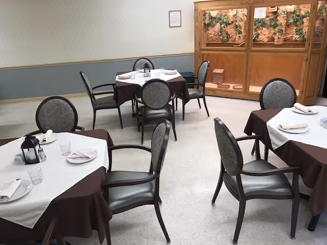 Small dining room with round tables covered in white and brown tablecloths and empty chairs arranged around them.