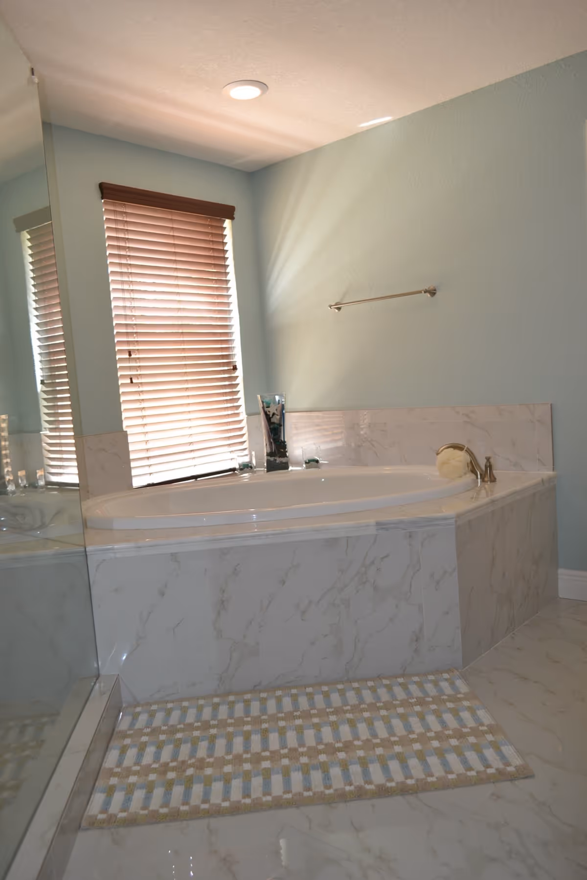 A bathroom featuring a large white marble bathtub with a silver faucet, a window with closed wooden blinds, a towel rack on the wall, and a patterned bath mat on the marble floor.