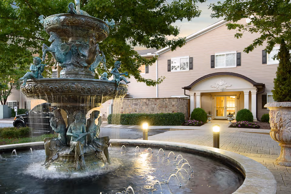 A decorative multi-tiered fountain with statues of musicians and horses in a circular pool, situated in front of the entrance to a beige building with black shutters and a covered porch. The area is landscaped with bushes, flowers, and trees, and there are two lit pathway lights near the entrance.