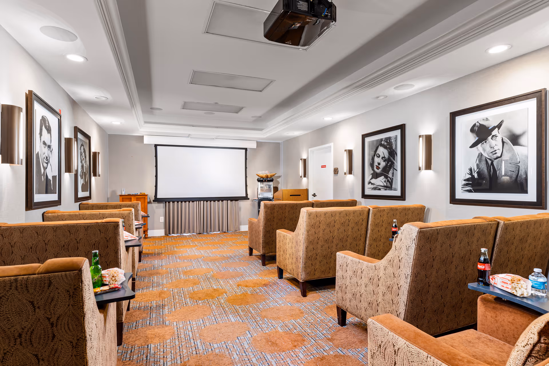 Small movie screening room with upholstered armchairs facing a projection screen and framed black-and-white portraits on the walls.