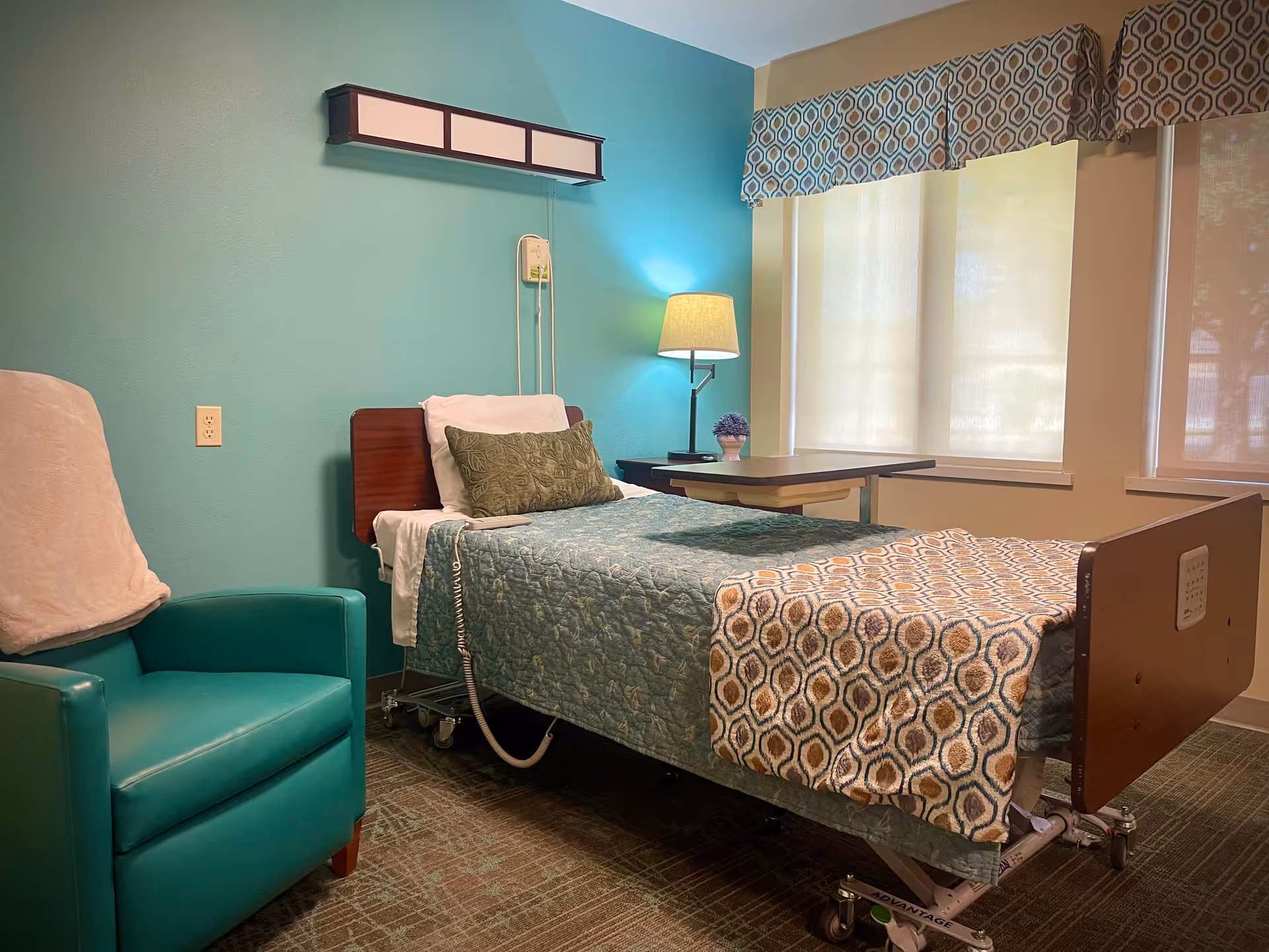 A neatly arranged room in a retirement facility featuring a hospital-style bed with blue and patterned bedding, a green armchair with a white cover, a bedside table with a lamp and a small plant, and windows with patterned valances and blinds.