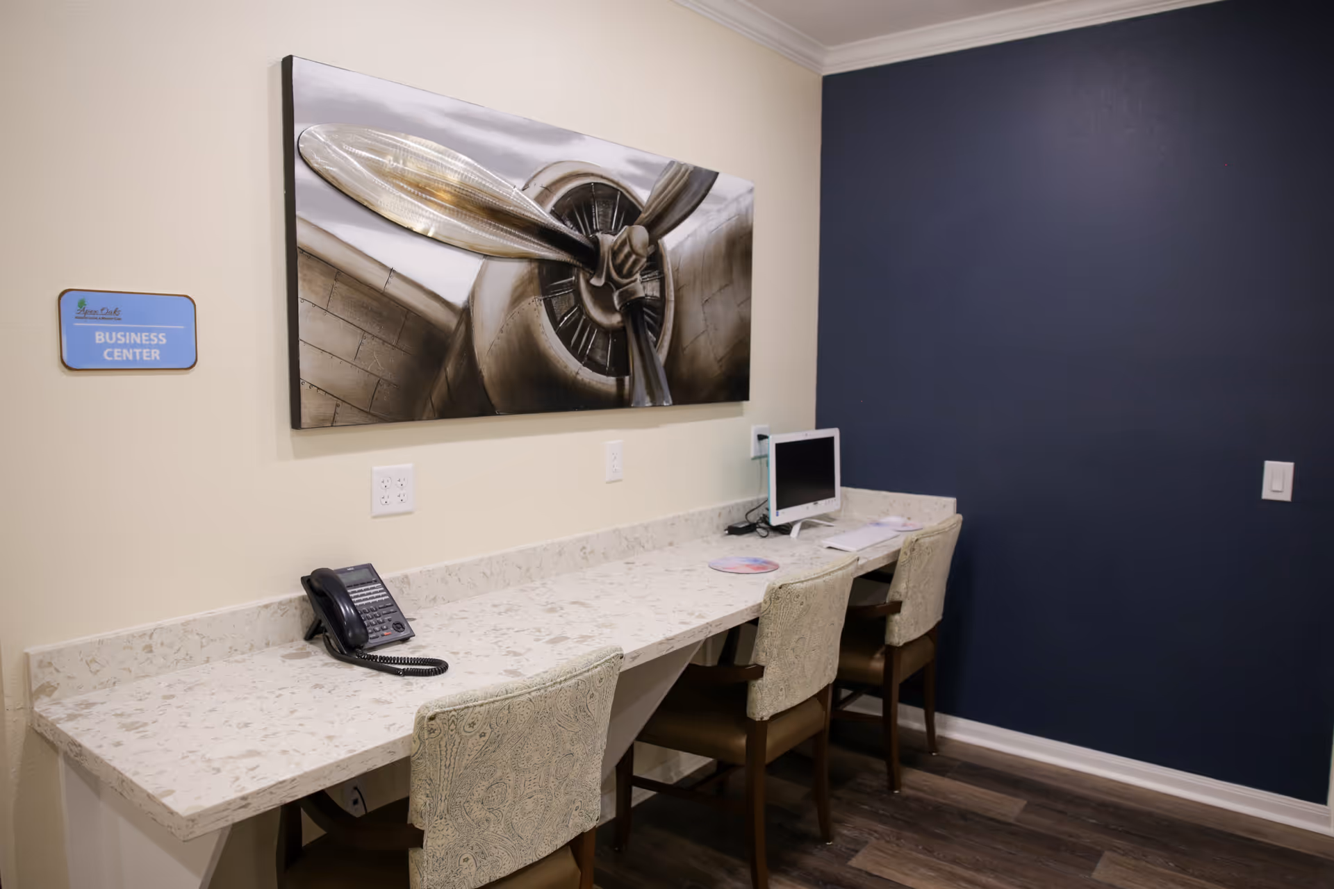 A business center area with a long marble countertop, three upholstered chairs, a telephone, and a computer. A large black and white photo of an airplane propeller is mounted on the wall above the counter. The wall to the right is painted dark blue, and there is a sign on the left wall that reads 'Apex Oaks Business Center.'