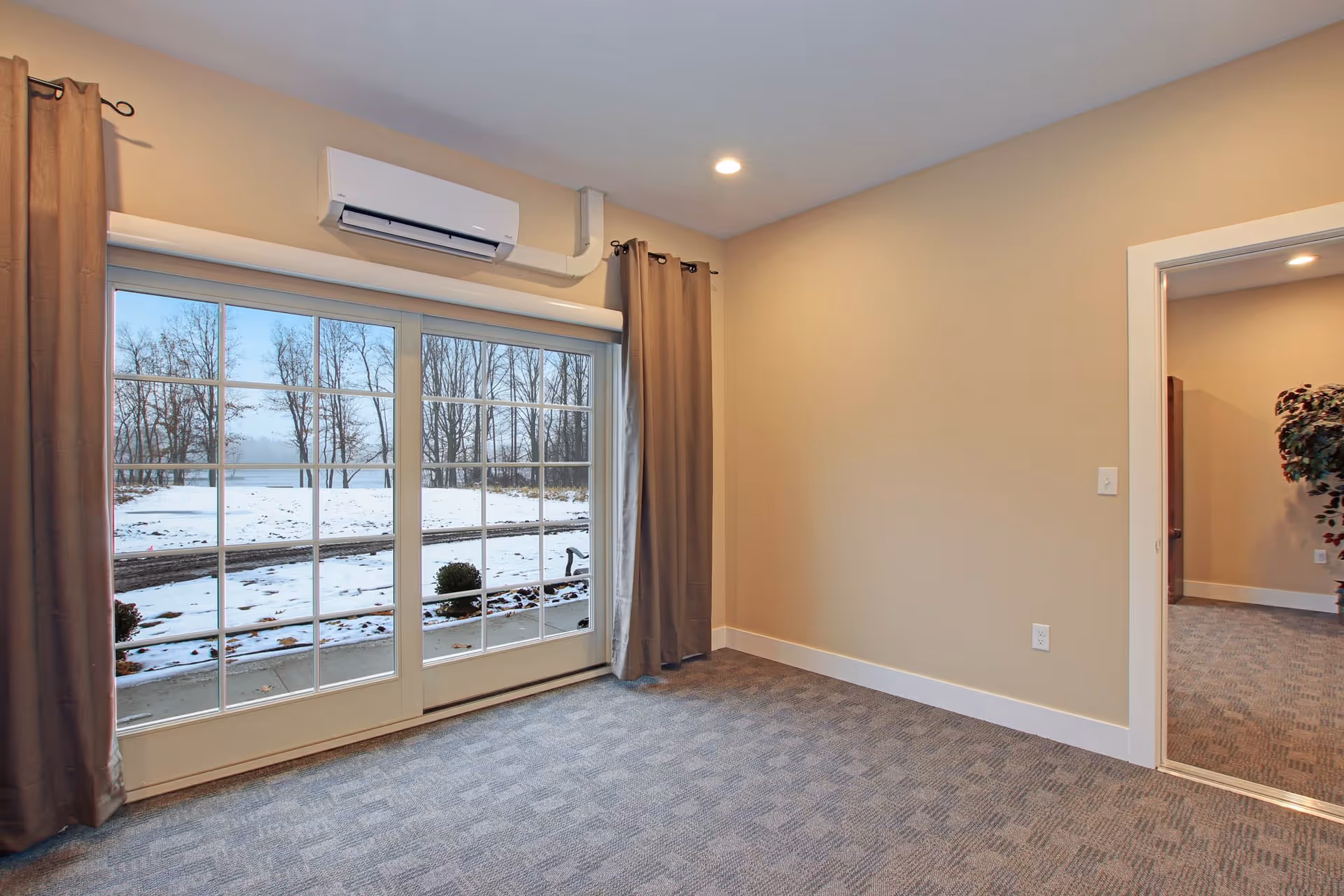 Empty room with beige walls, gray carpet, large sliding glass doors with brown curtains, and a view of a snowy outdoor landscape with bare trees. A doorway leads to another room with similar carpeting and a potted plant visible.