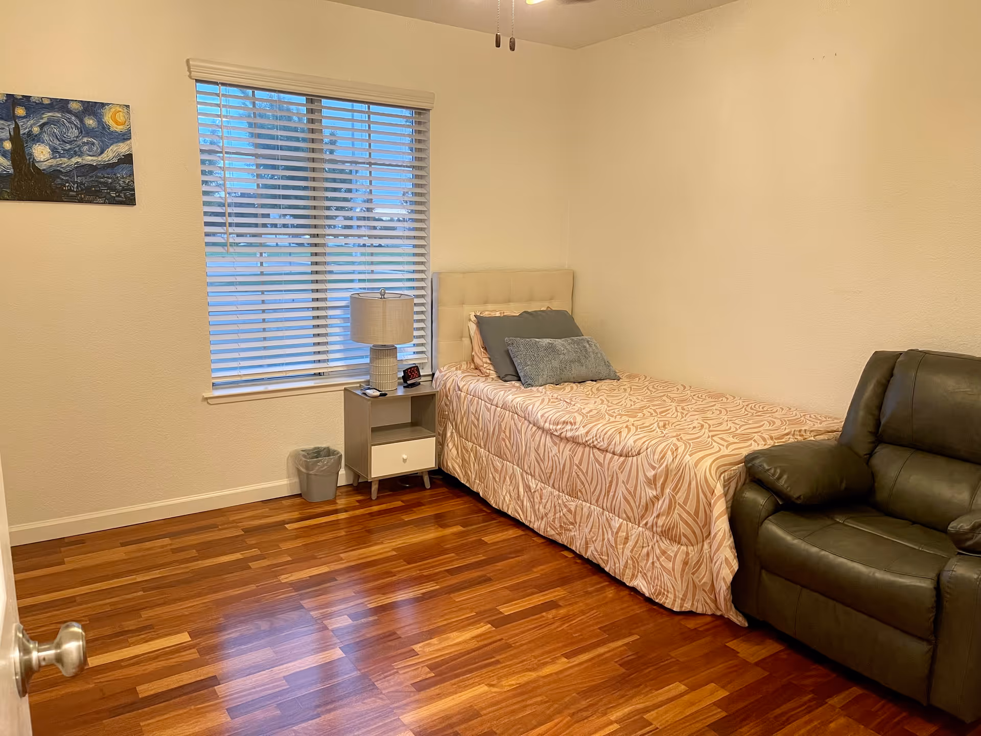 A small bedroom with a single bed covered in a patterned beige bedspread and several pillows. Next to the bed is a nightstand with a lamp, a clock, and a small item on it. There is a window with white blinds behind the nightstand. A dark green recliner chair is positioned to the right of the bed. The floor is wooden, and a small painting resembling Van Gogh's Starry Night hangs on the wall to the left of the window.